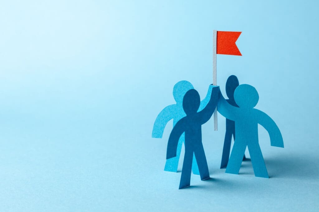Three blue paper-cut figures standing together and raising a red flag against a light blue background, symbolizing teamwork and leadership.